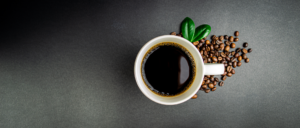Photo shows a cup of black coffee with coffee beans and green leaves around the base of the cup.