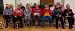 Photo shows the choir seated in rows, wearing festive jumpers and hats, song sheets in hand.